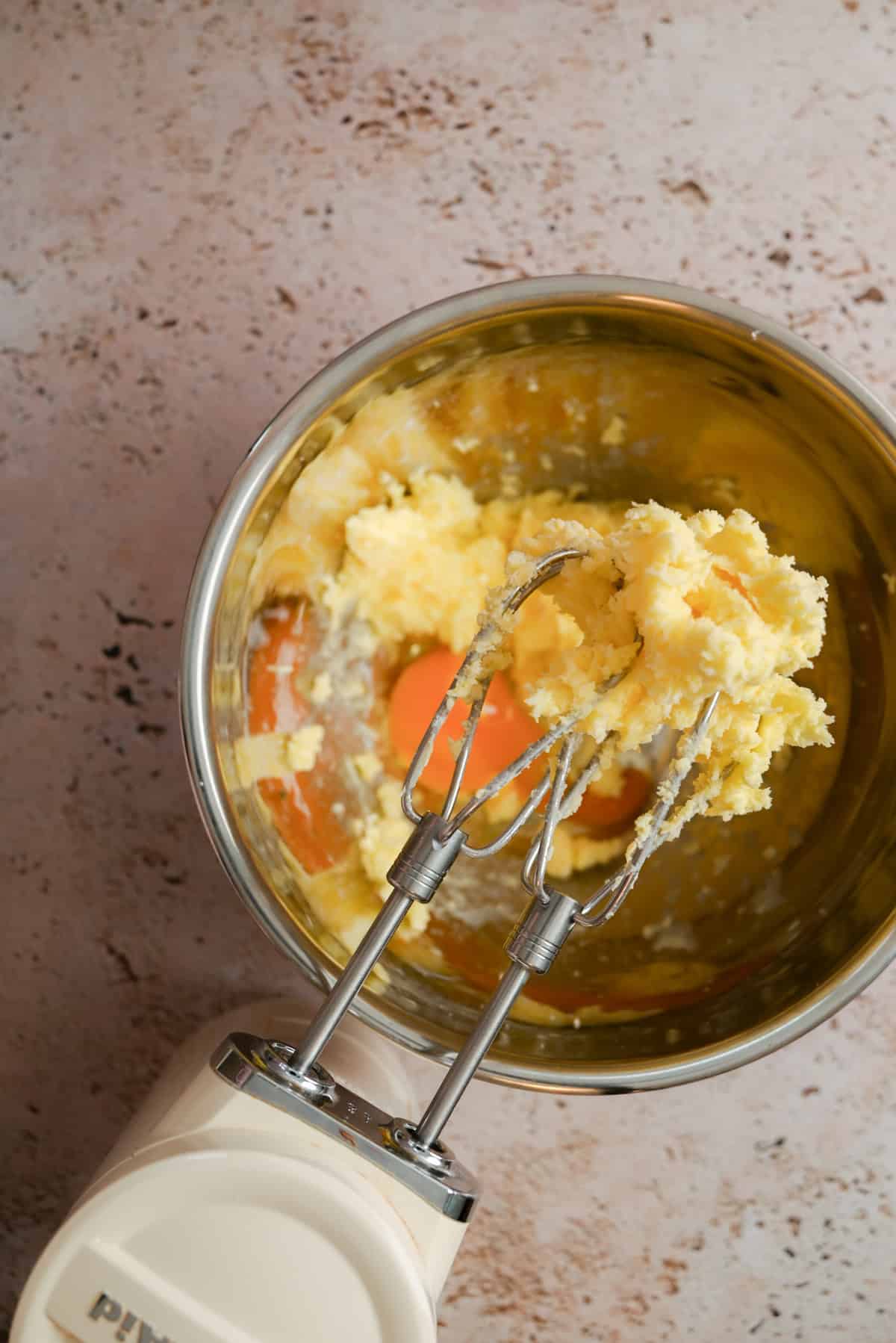 A mixing bowl containing creamed butter and sugar with an egg, ready for a Cranberry Meringue Tart, and an electric hand mixer with beaters covered in the mixture resting on the edge of the bowl.