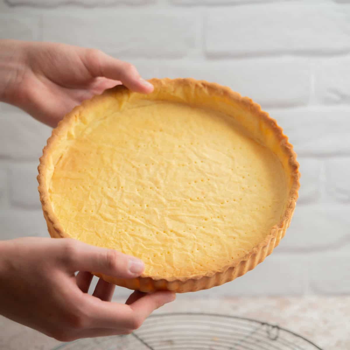 A person holds a freshly baked Pâte Sucree tart crust with a golden, flaky edge, ready to be filled. The background features a white brick wall.