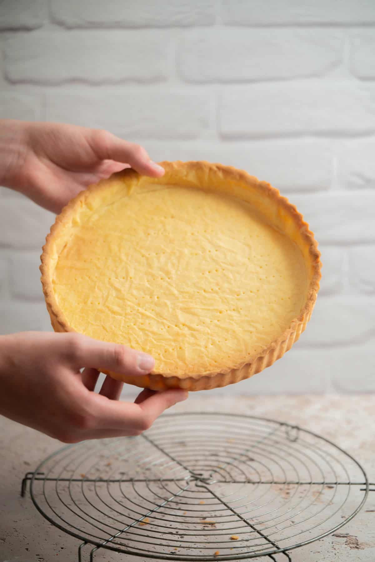 Two hands hold a freshly baked, empty pâte sucrée pie crust above a cooling rack on a light surface, with a white brick wall background.