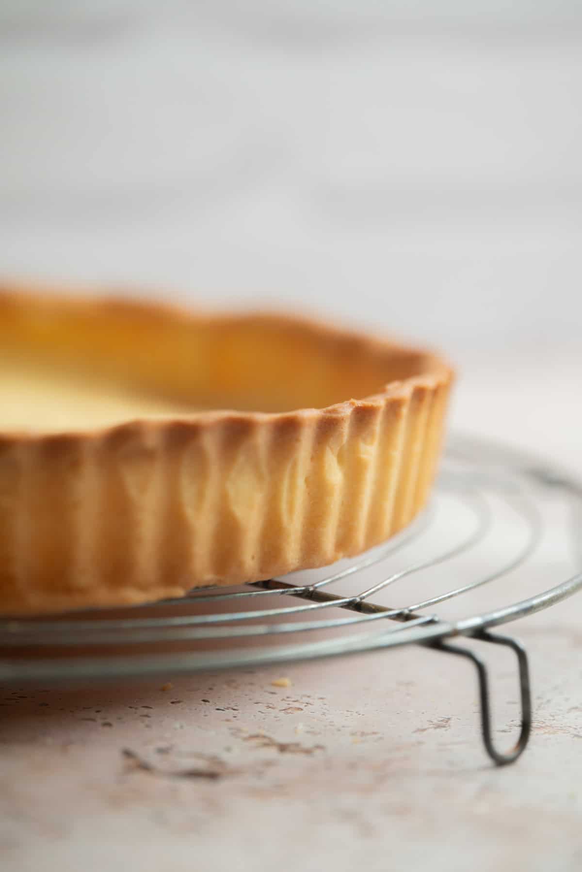 A close-up of a golden-brown pâte sablée tart crust resting on a round metal cooling rack, with a softly blurred background.