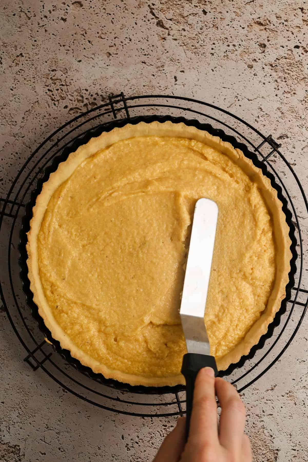 A hand uses an offset spatula to spread a creamy filling evenly inside a tart crust on a round wire cooling rack, set against a textured, light brown surface.