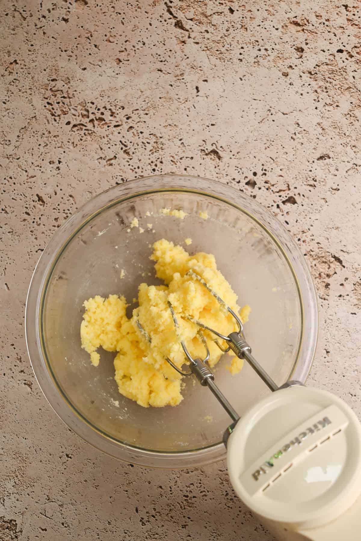 A glass bowl with yellow creamed butter and sugar sits on a textured surface; a handheld electric mixer with beaters is partially visible and resting inside the bowl.
