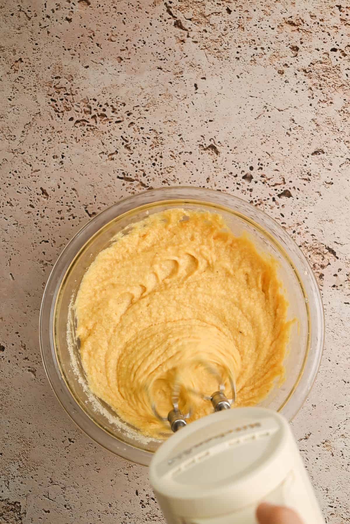 A hand holds an electric mixer blending yellow batter in a clear glass bowl on a textured countertop.
