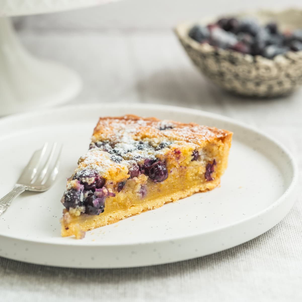 A slice of blueberry frangipane tart dusted with powdered sugar sits on a white plate next to a fork, with a bowl of fresh blueberries blurred in the background.