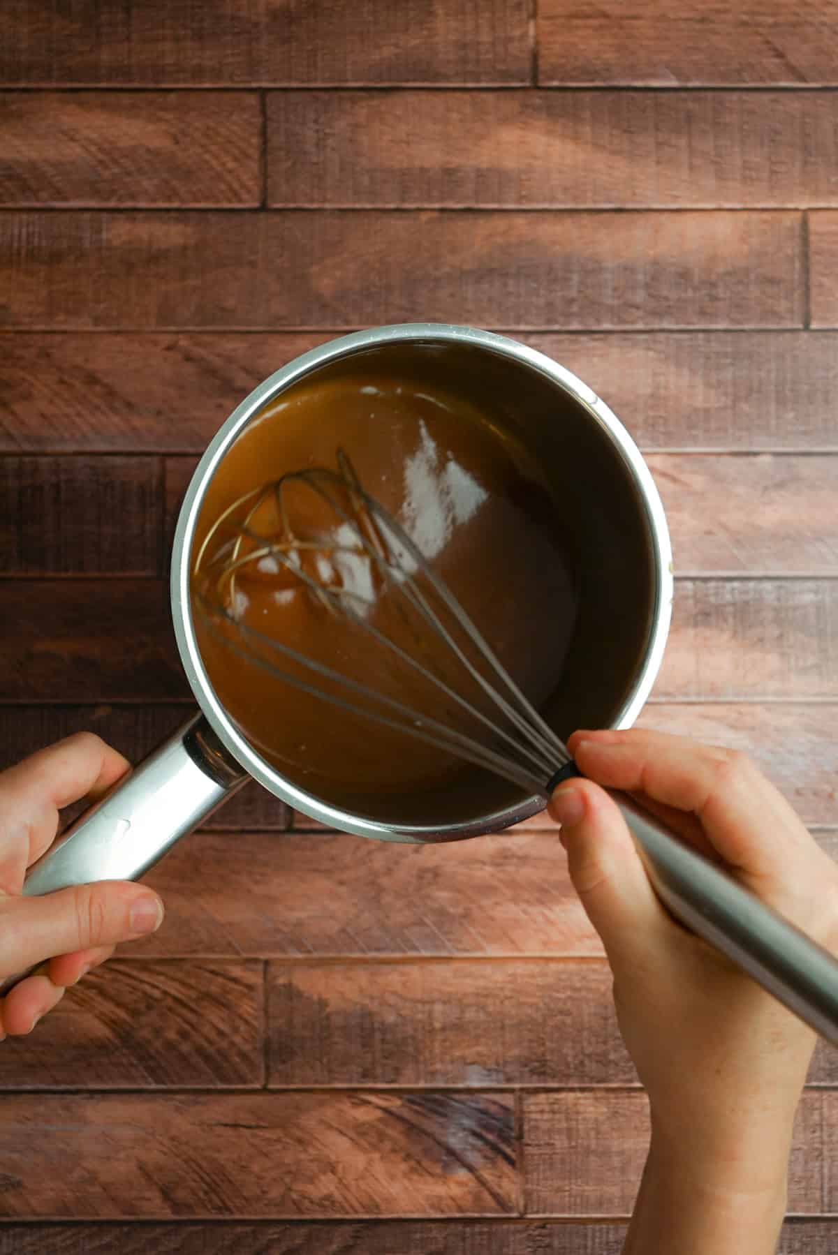 A person holds a saucepan of caramel sauce and stirs it with a metal whisk on a wooden surface.