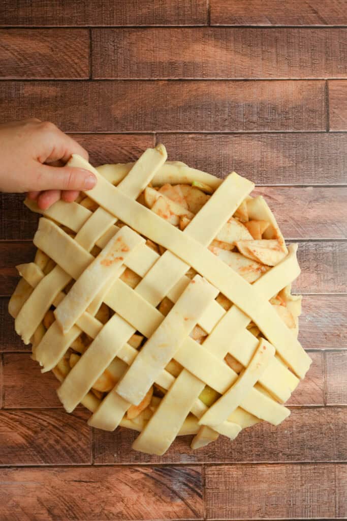 A hand is weaving strips of pie dough in a lattice pattern over an unbaked apple pie on a wooden surface.