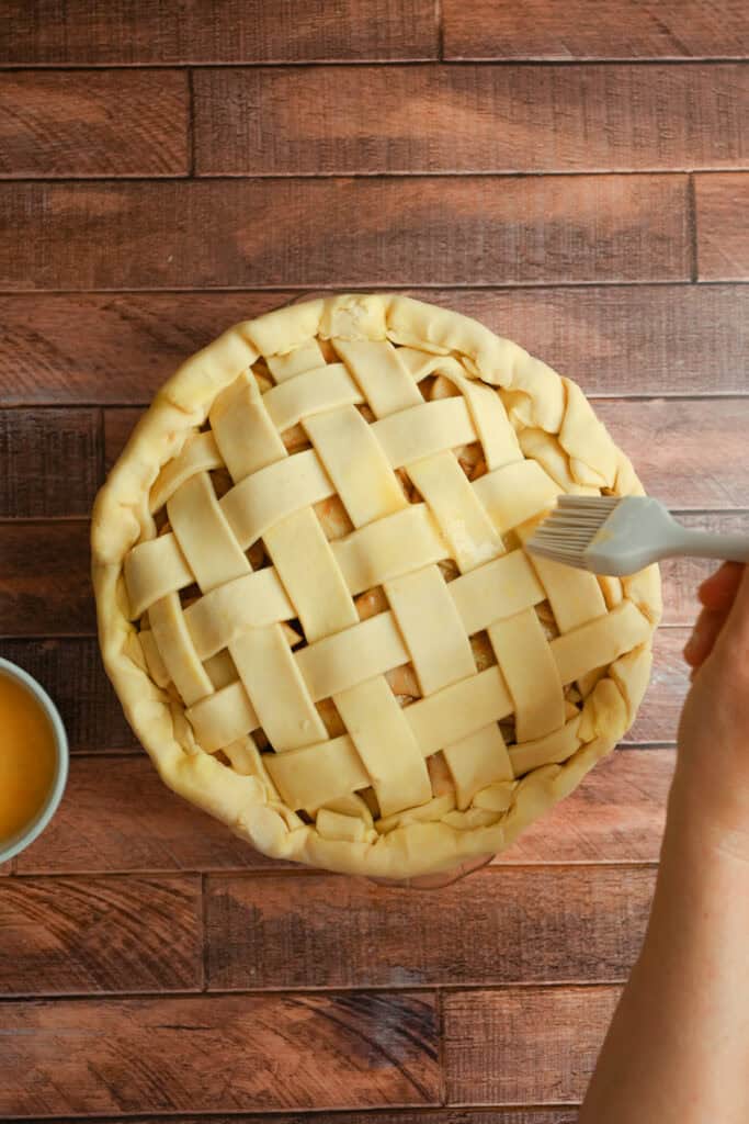 A hand uses a brush to apply egg wash to a pie with a lattice crust on a wooden surface. A small bowl of egg wash sits nearby.