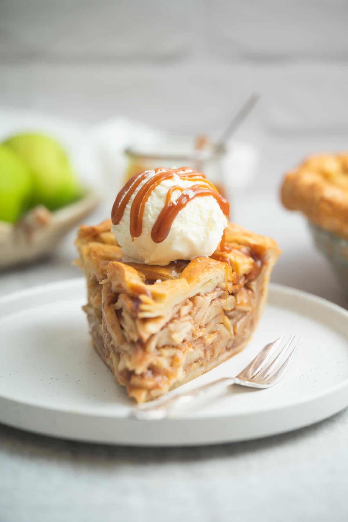 A slice of salted caramel apple pie topped with a scoop of vanilla ice cream and caramel drizzle sits on a white plate with a fork; green apples and another pie are blurred in the background.