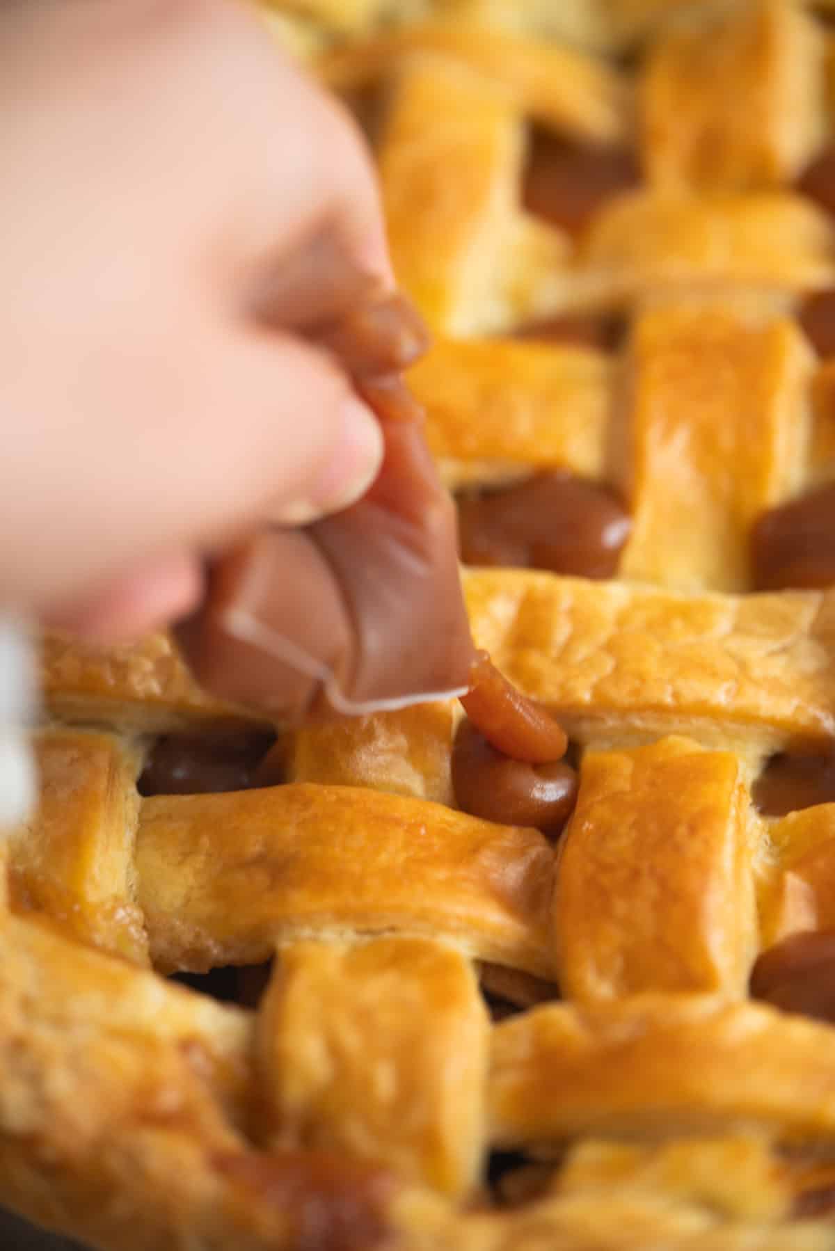 A close-up of a hand placing or adjusting a piece of caramel on top of a golden-brown lattice-topped pie.