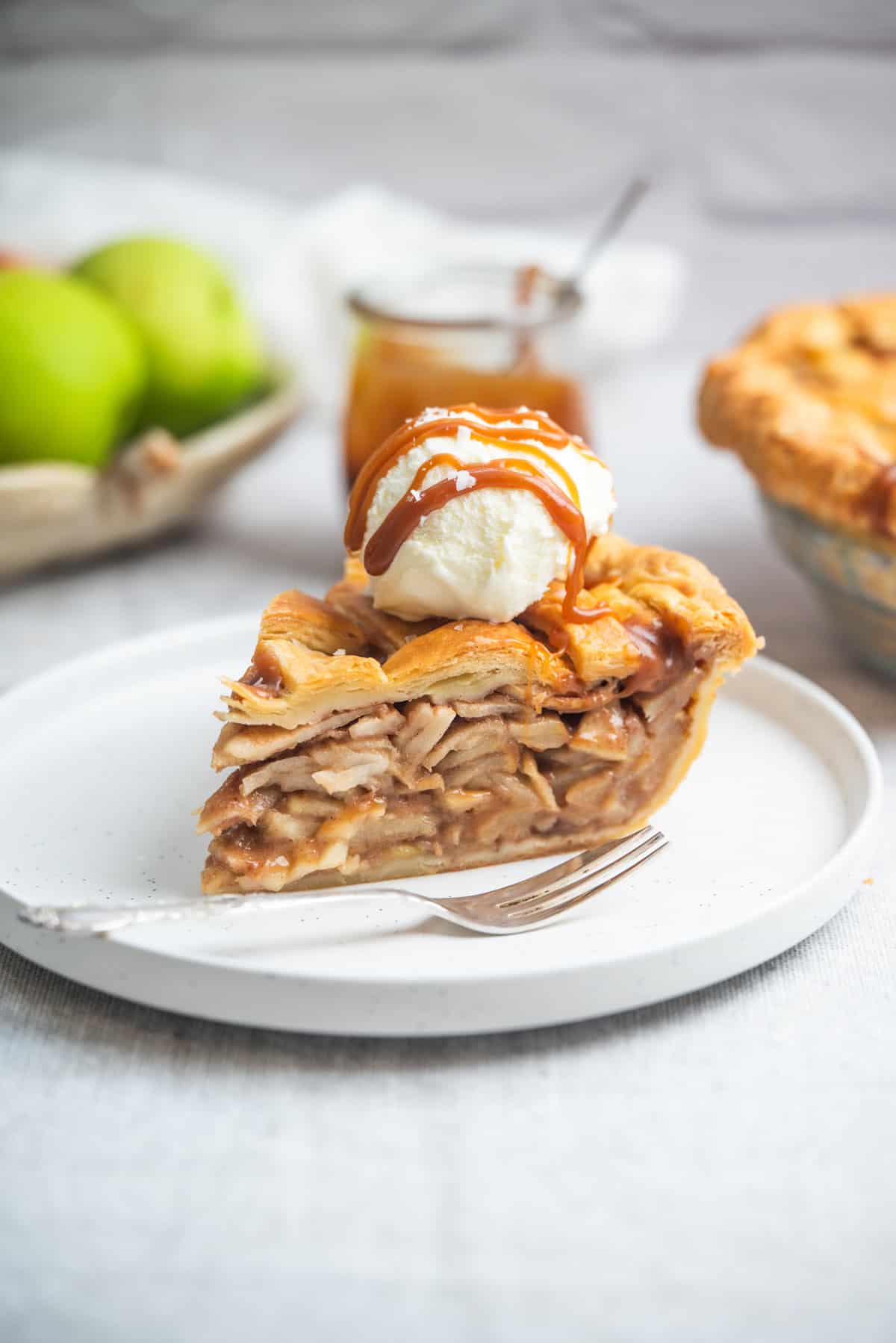 A slice of apple pie topped with a scoop of vanilla ice cream and caramel drizzle sits on a white plate with a fork. There are green apples and a jar of caramel sauce in the background.