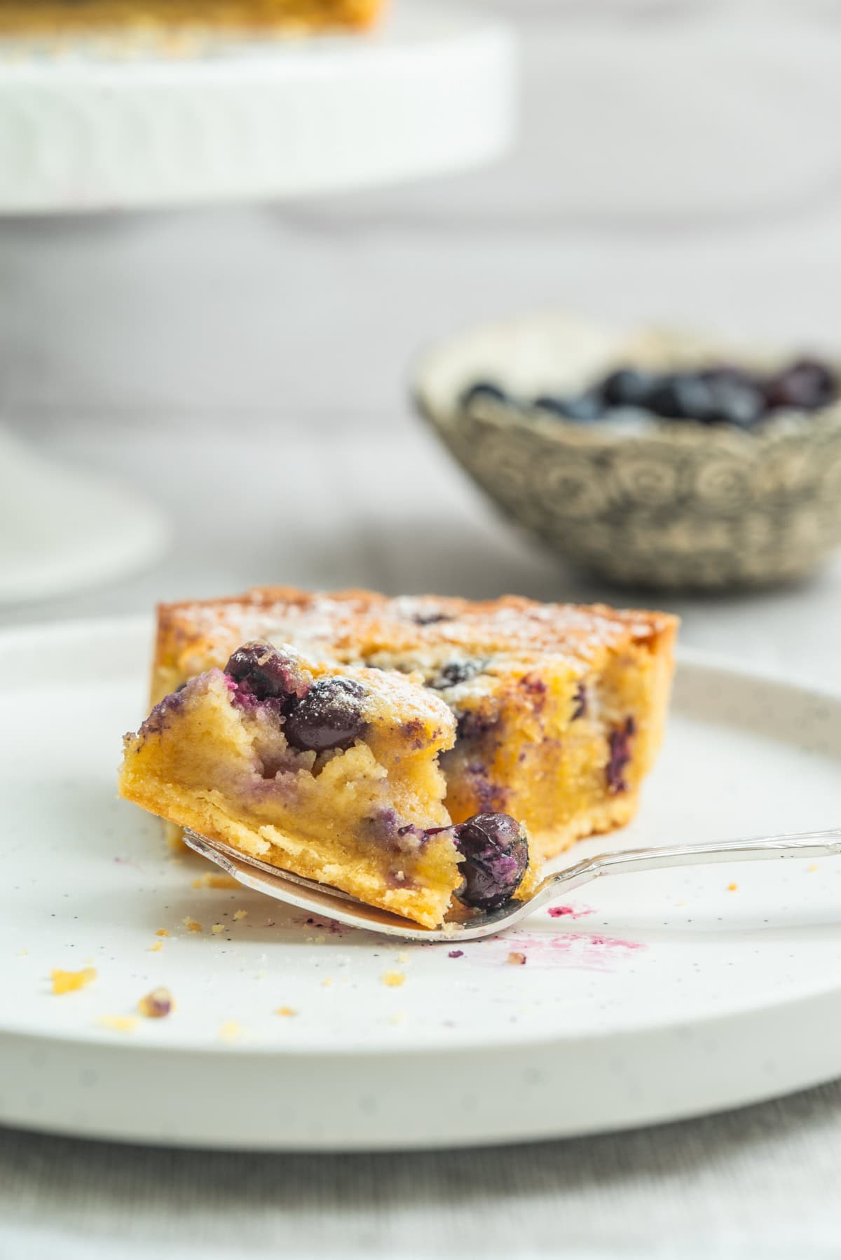 A slice of blueberry cake on a white plate with a fork holding a bite, blurred bowl of blueberries in the background.