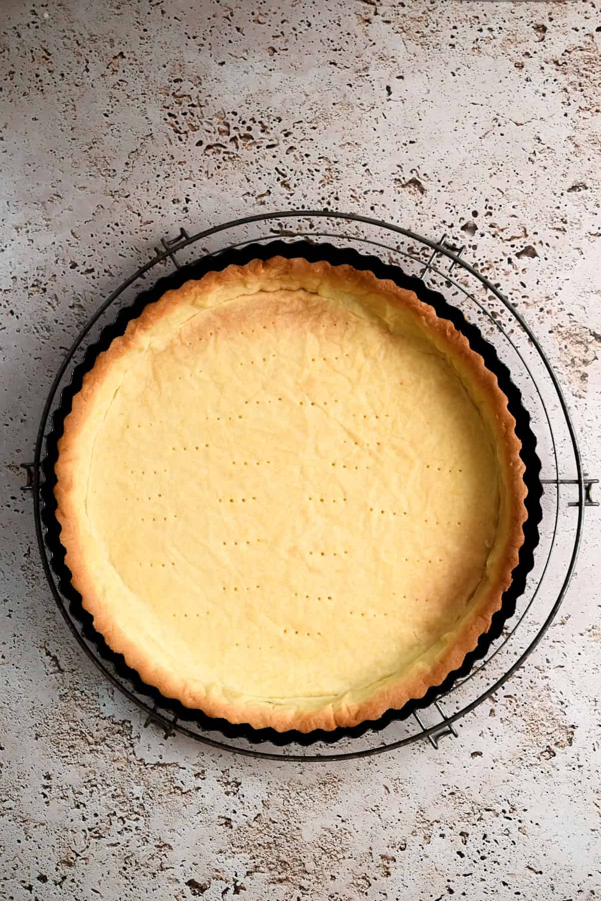 A baked pâte sucrée tart crust in a round pan sits on a wire cooling rack over a textured light-colored surface. The golden brown crust features tiny fork holes visible on the bottom.