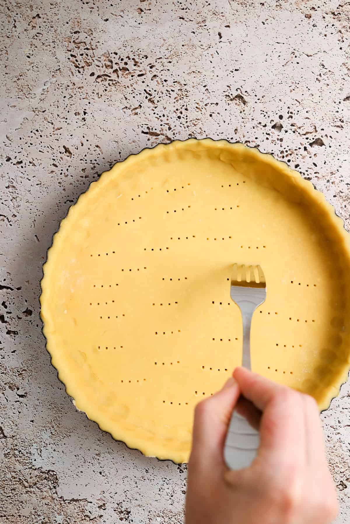 A hand is using a fork to prick holes in an unbaked crust in a round tart pan, set on a textured, light-colored surface.