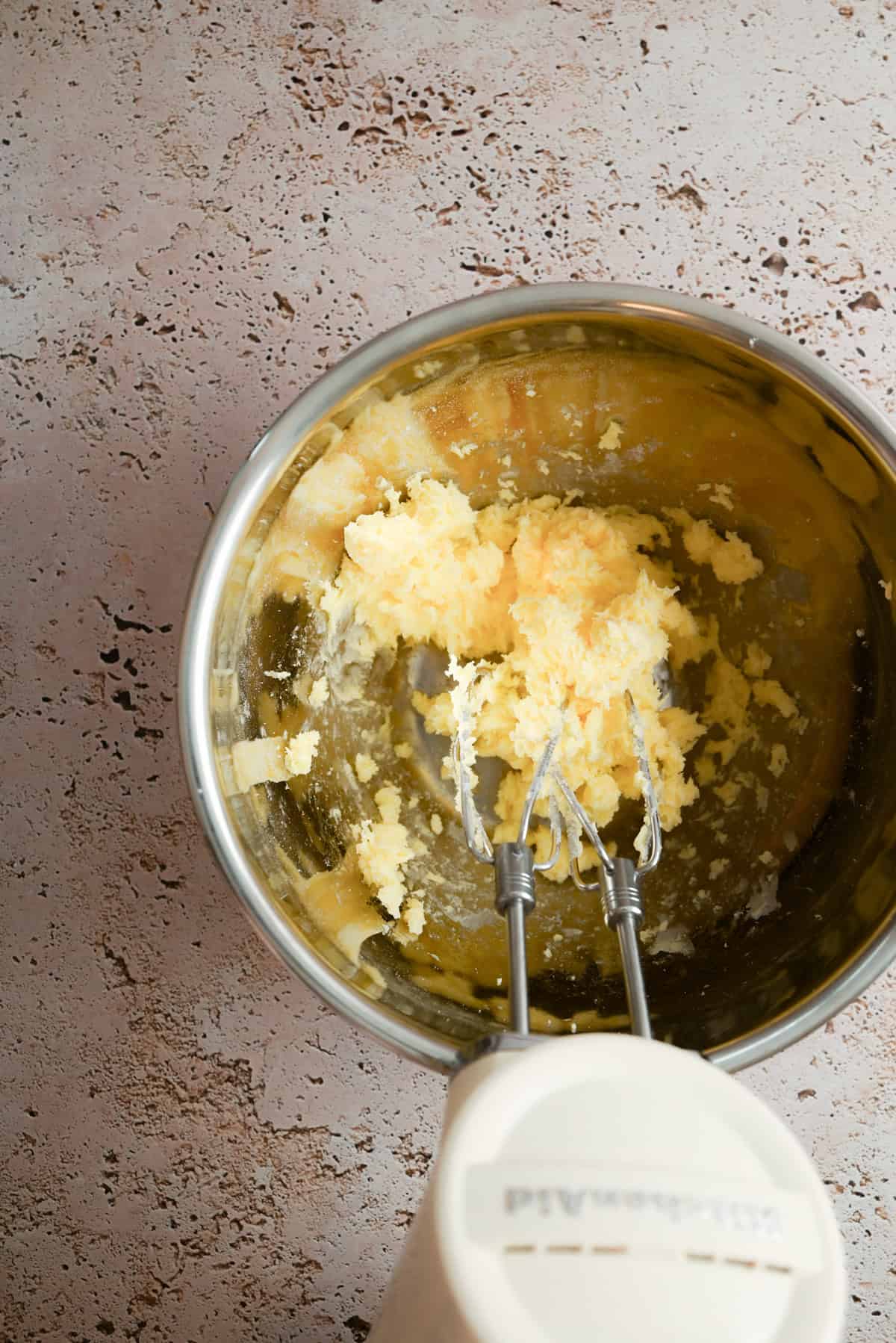 A hand mixer blends softened butter and sugar in a stainless steel bowl on a light textured countertop.