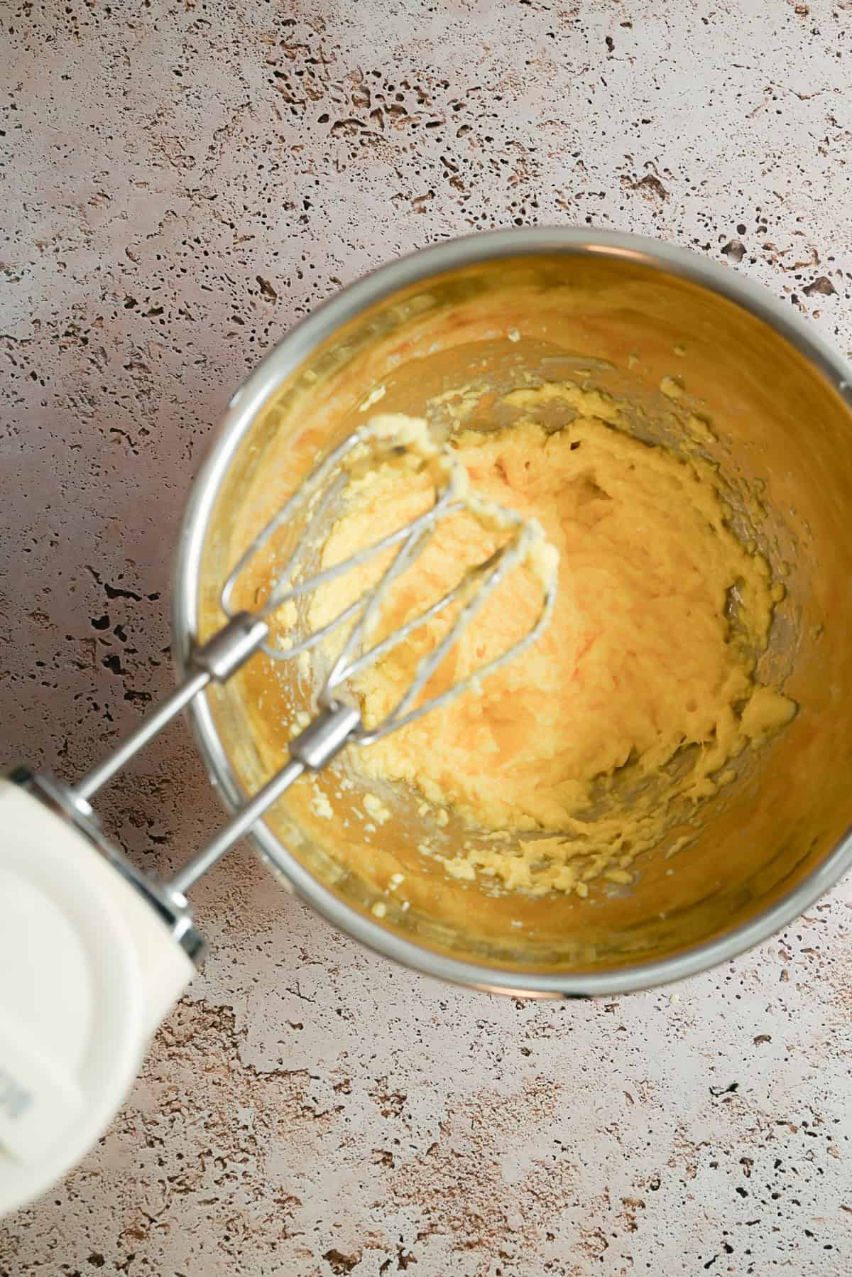 A metal mixing bowl with creamy yellow batter being mixed by an electric hand mixer, placed on a textured light brown surface.