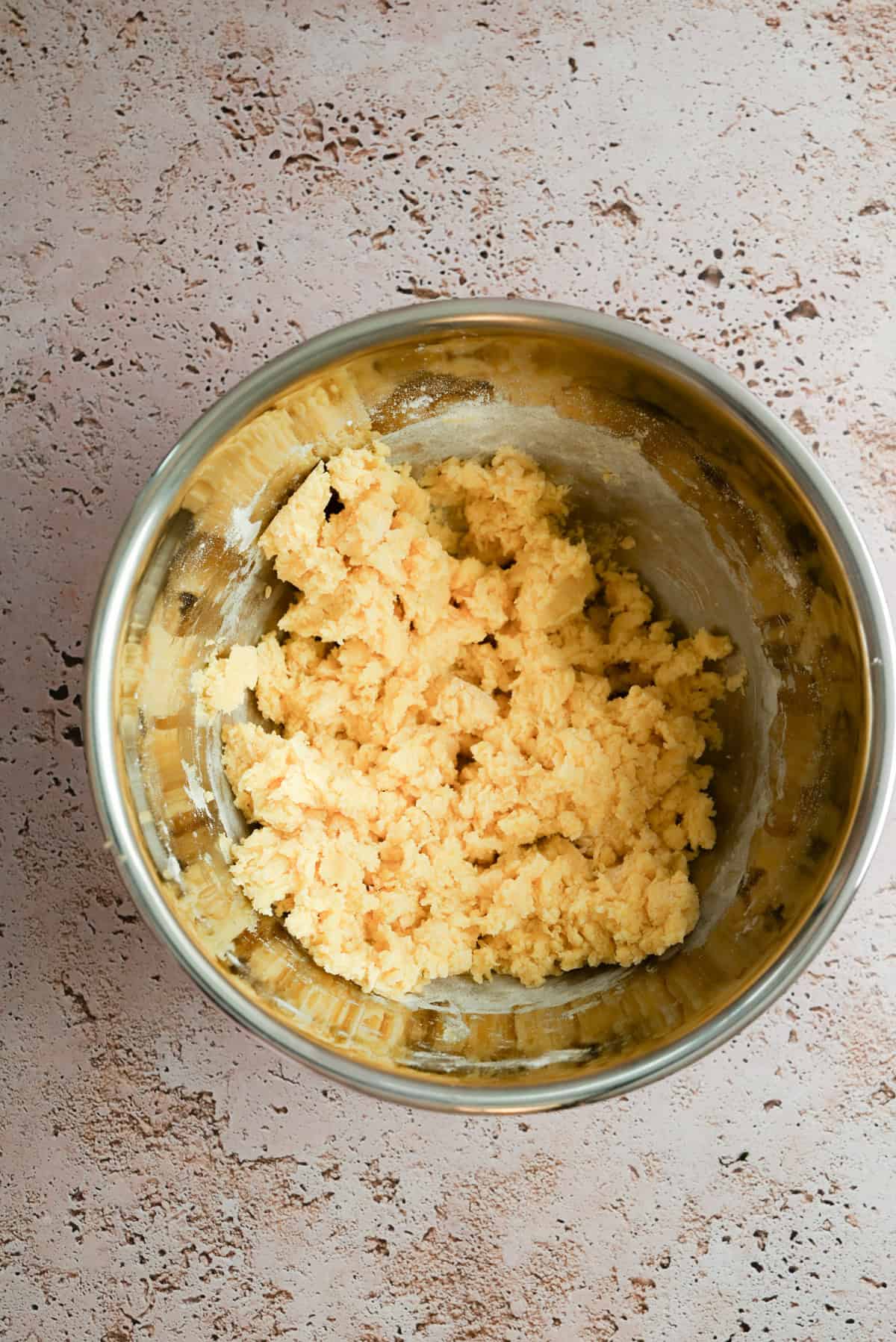 A metal mixing bowl containing pale yellow dough sits on a textured light brown countertop.