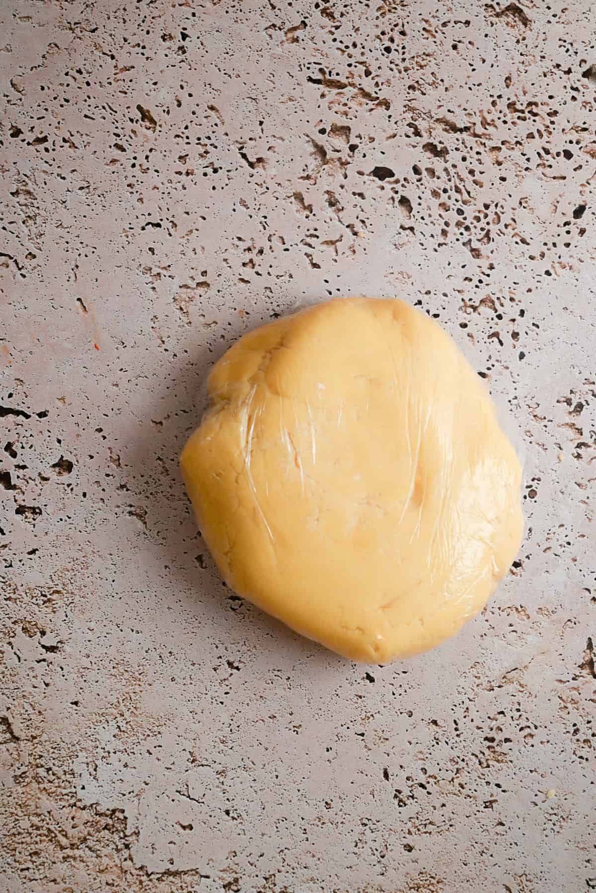 A round disk yellow dough wrapped in plastic sits on a textured, light-colored surface.