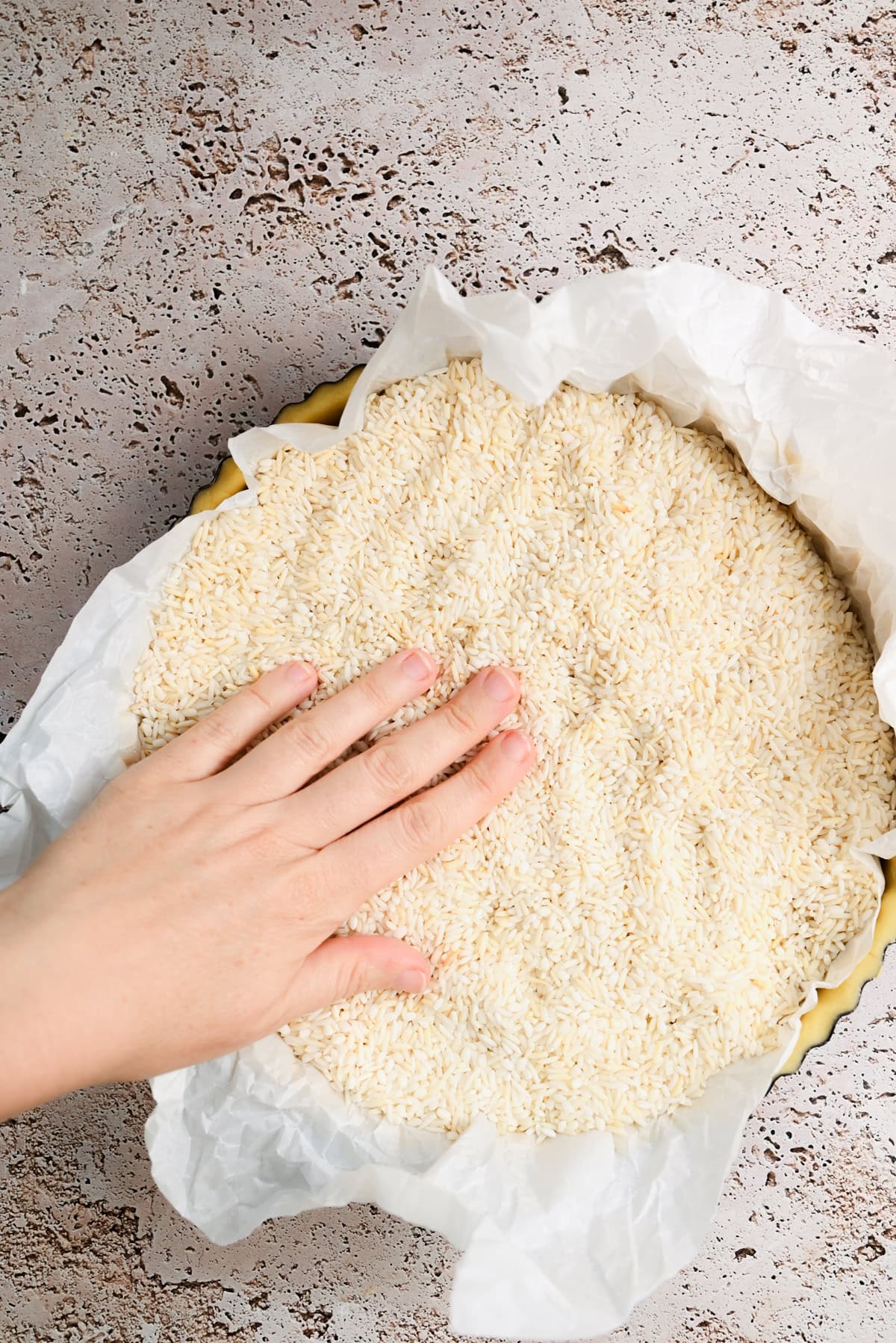 A hand presses down on uncooked rice over parchment paper in a round tart pan