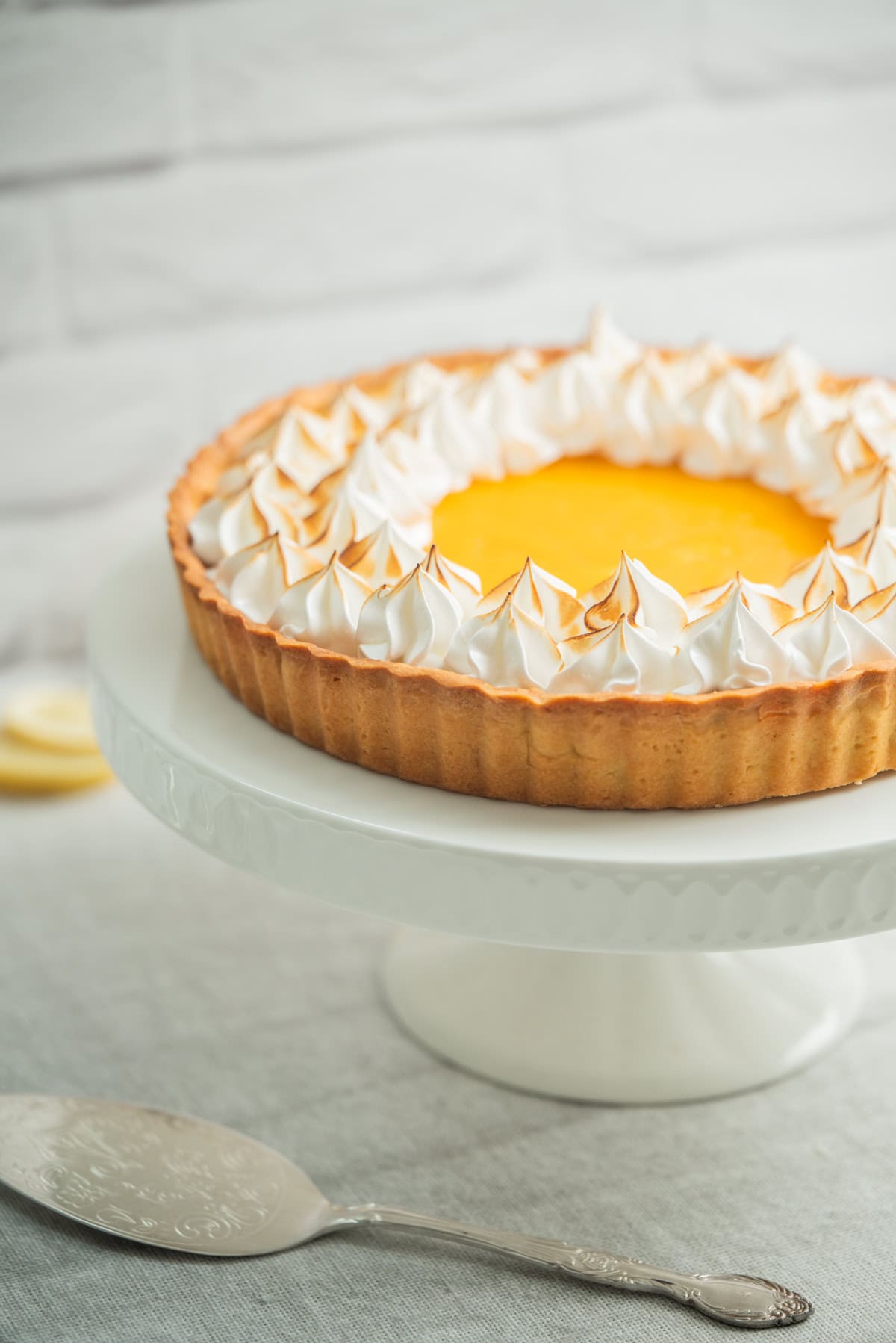 A lemon meringue tart with golden brown peaks of meringue sits on a white cake stand, with a serving utensil nearby on a light gray surface.