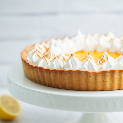 A lemon meringue tart with golden-brown peaks of meringue sits on a white cake stand. A halved lemon is blurred in the background.