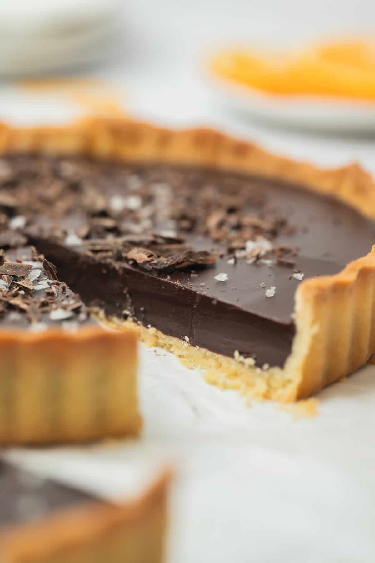 A close-up of a chocolate tart with a flaky crust, topped with chocolate shavings and sea salt. A slice has been removed, revealing the smooth, rich chocolate filling inside.