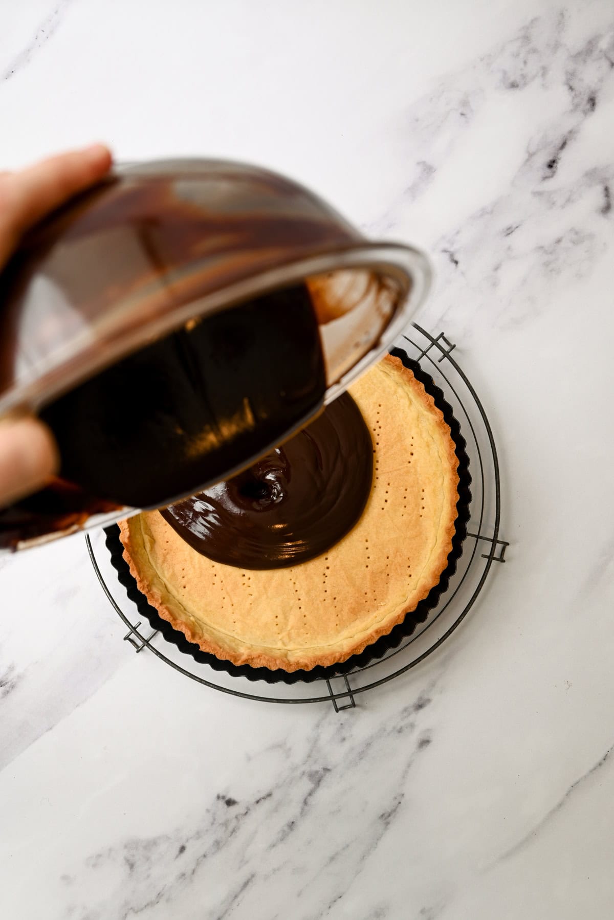 A hand pours glossy melted chocolate from a glass bowl into a baked tart crust sitting on a wire rack over a marble surface.