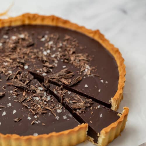 A chocolate tart with a golden crust, topped with chocolate shavings and sea salt, has one slice slightly pulled out. In the background, a plate with orange slices is blurred. The tart is on a white surface.