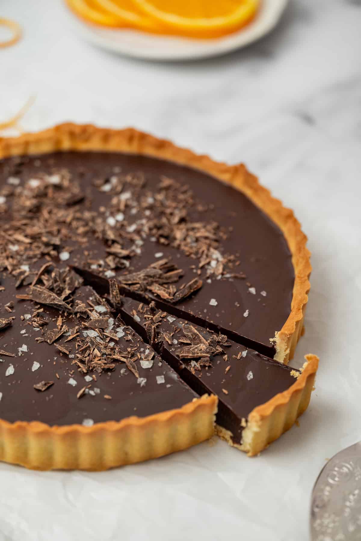 A chocolate tart with a golden crust, topped with chocolate shavings and sea salt, has one slice slightly pulled out. In the background, a plate with orange slices is blurred. The tart is on a white surface.