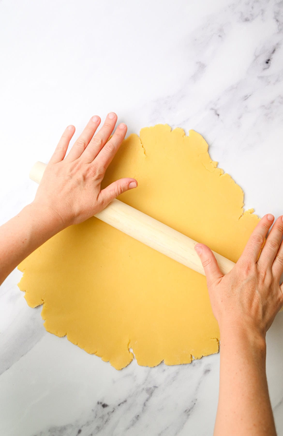 Hands rolling out yellow dough with a wooden rolling pin on a white marble surface.