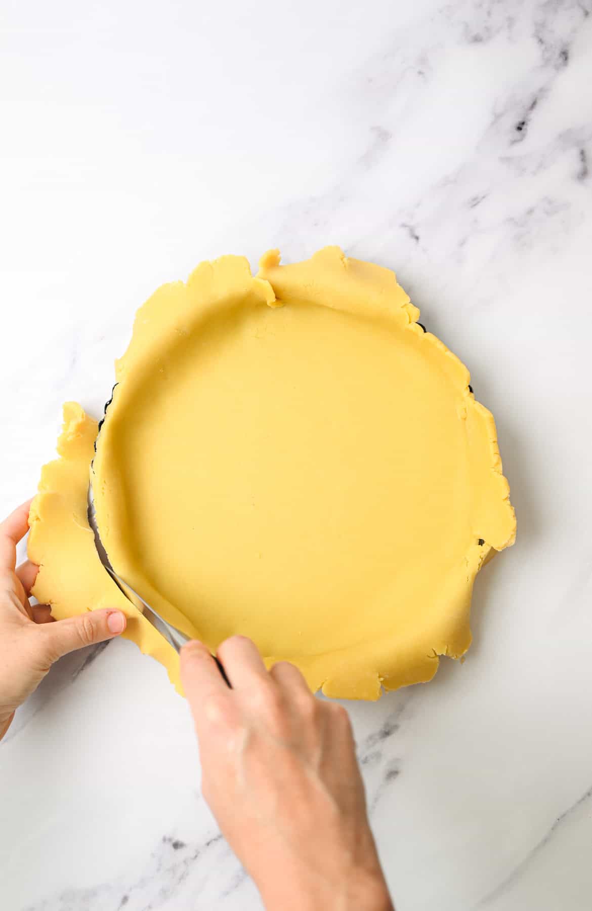 Hands trimming excess pie dough from the edge of a tart pan with a knife on a white marble surface.