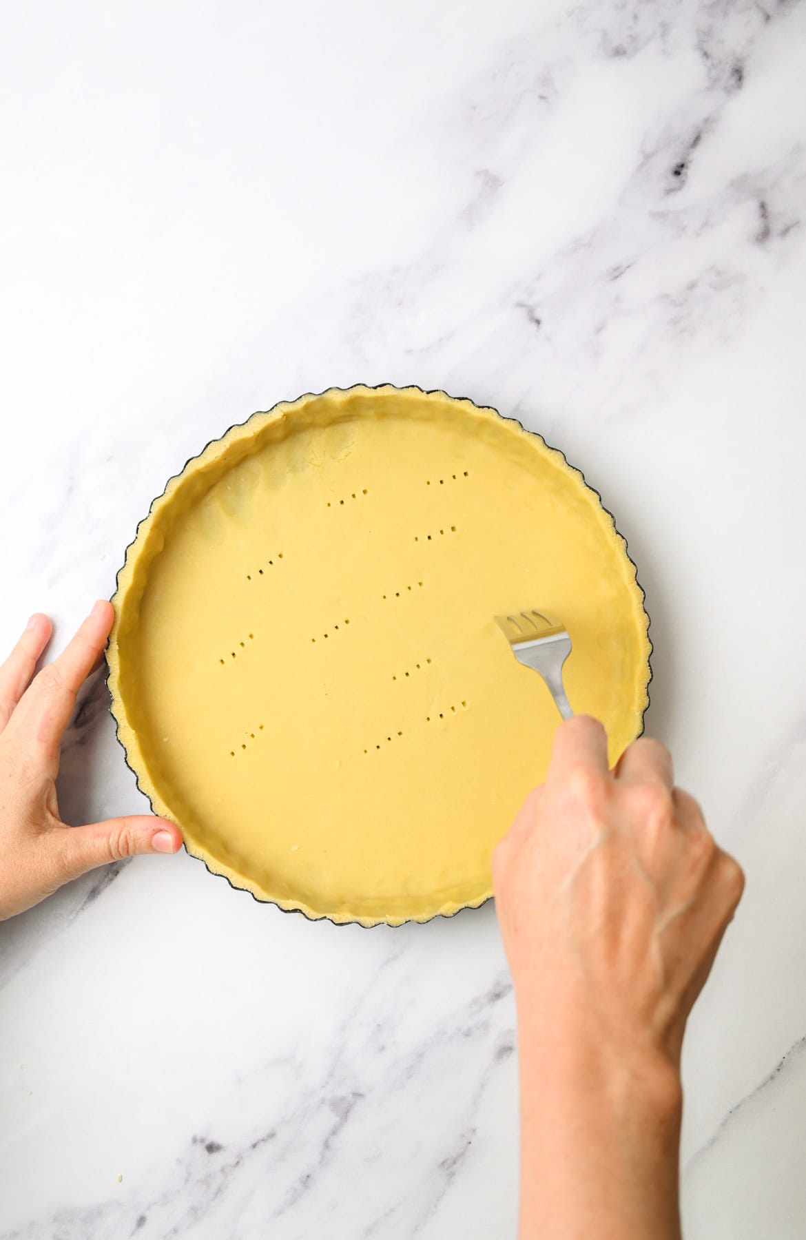 A person uses a fork to poke holes in the bottom of an unbaked pie crust in a round tart pan on a marble countertop.