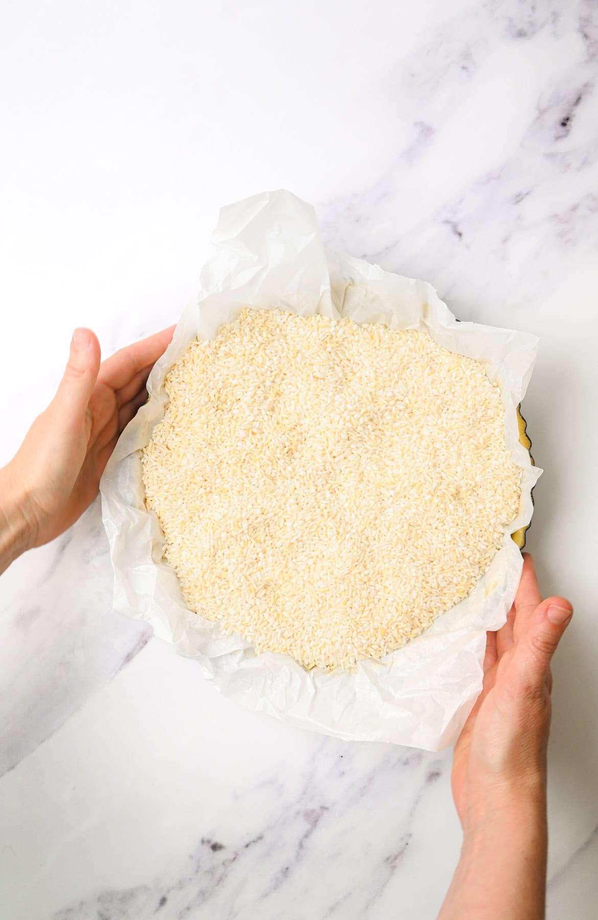 Two hands hold a round baking pan lined with crumpled parchment paper and filled with uncooked rice, likely being used for blind baking a pie crust. The scene is set on a white marble surface.