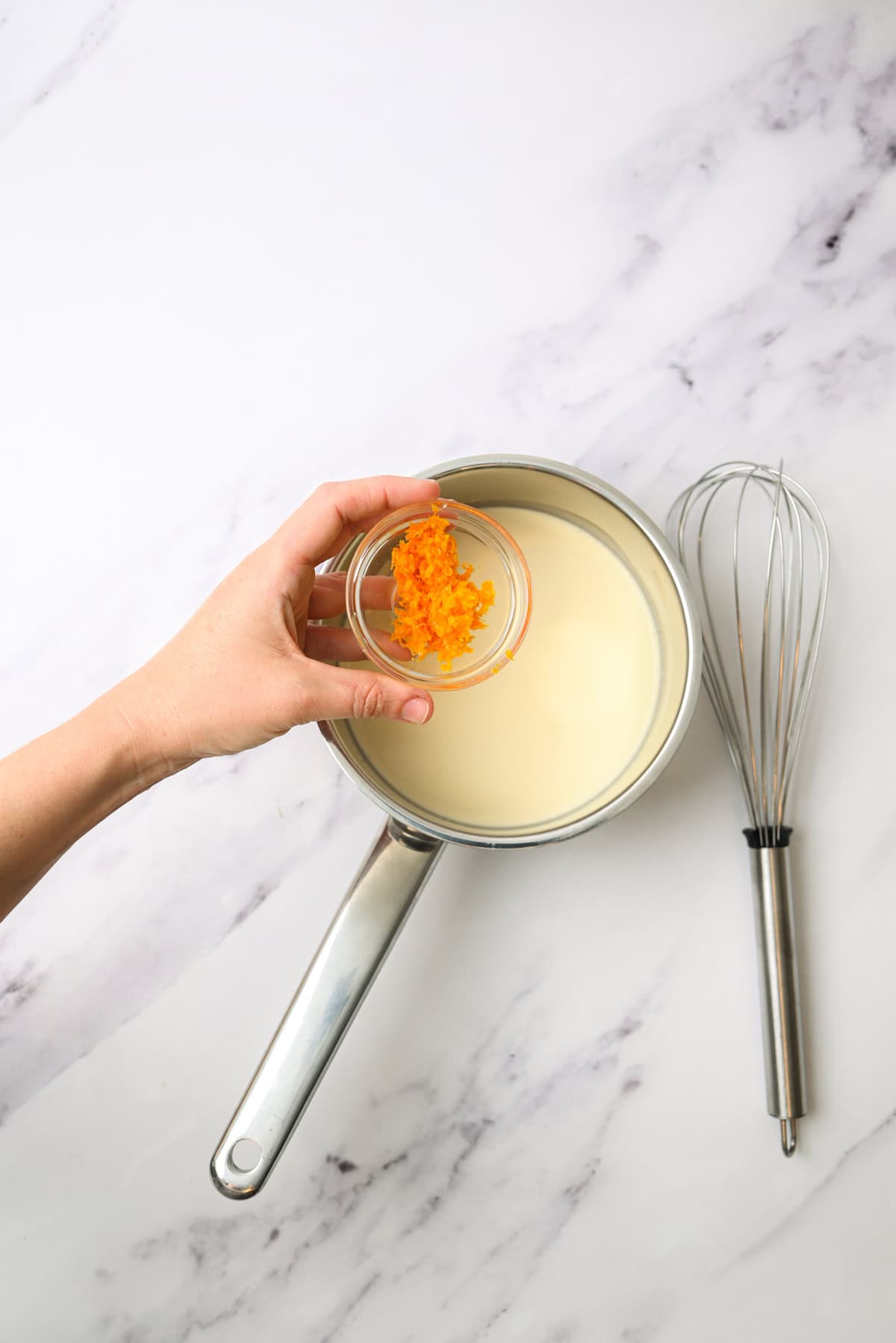 A hand holds a small bowl of orange zest over a saucepan filled with cream, next to a whisk on a white marble surface.