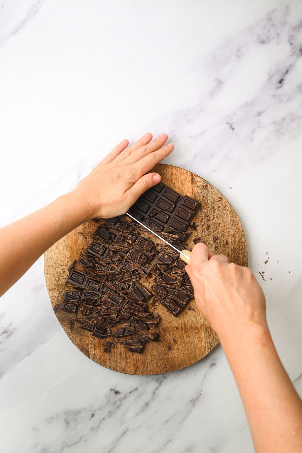 A person chops a bar of dark chocolate into small pieces on a round wooden cutting board placed on a white marble surface.