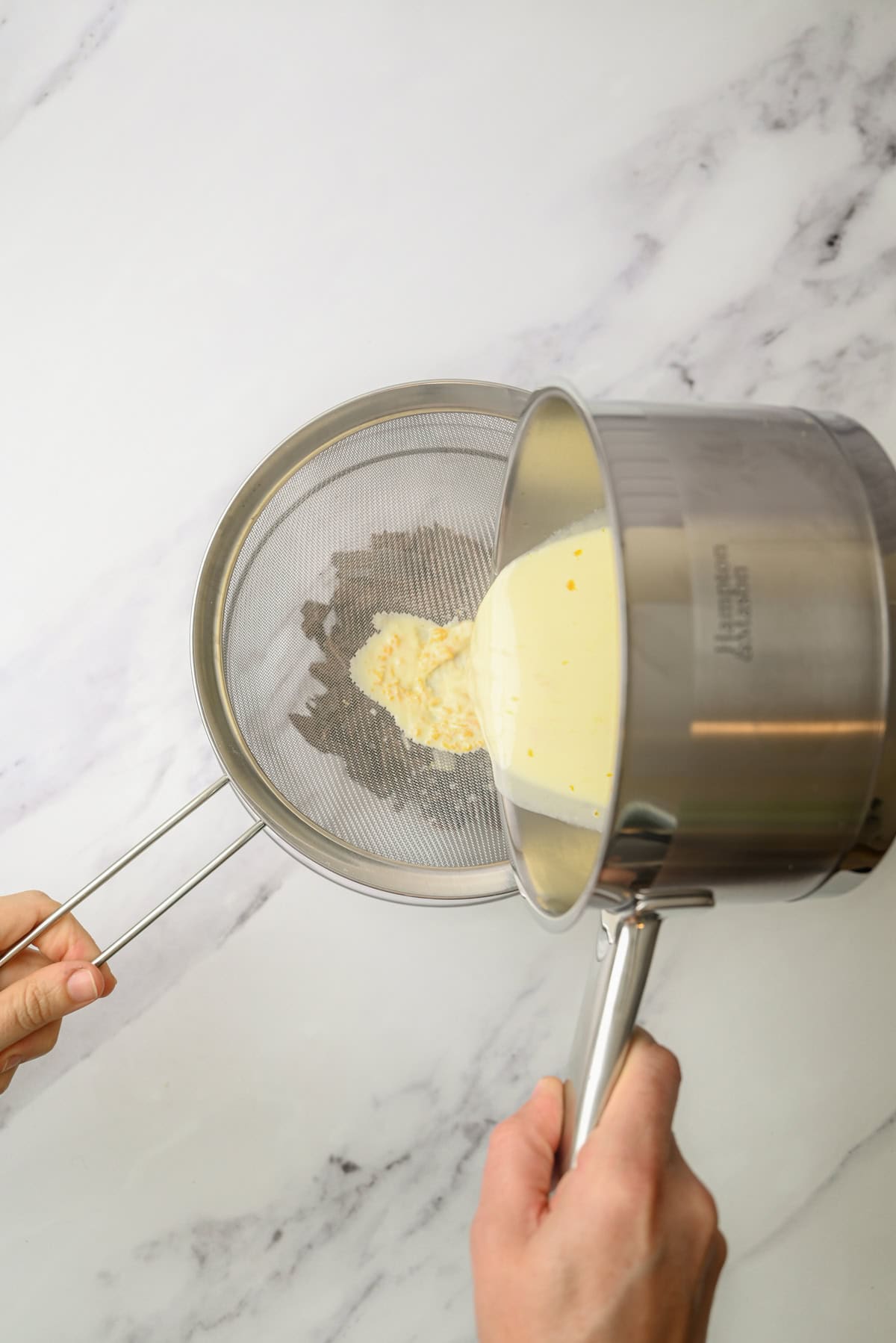 A person pours a creamy mixture from a saucepan through a fine mesh sieve, straining it over a white marble surface.