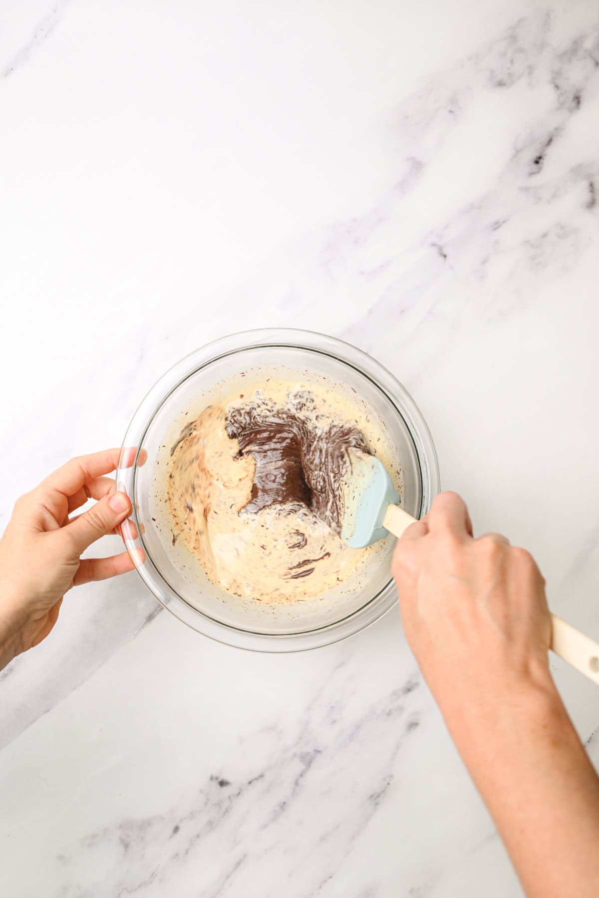 A person’s hands mixing a light batter with melted chocolate in a glass bowl using a spatula on a white marble surface.