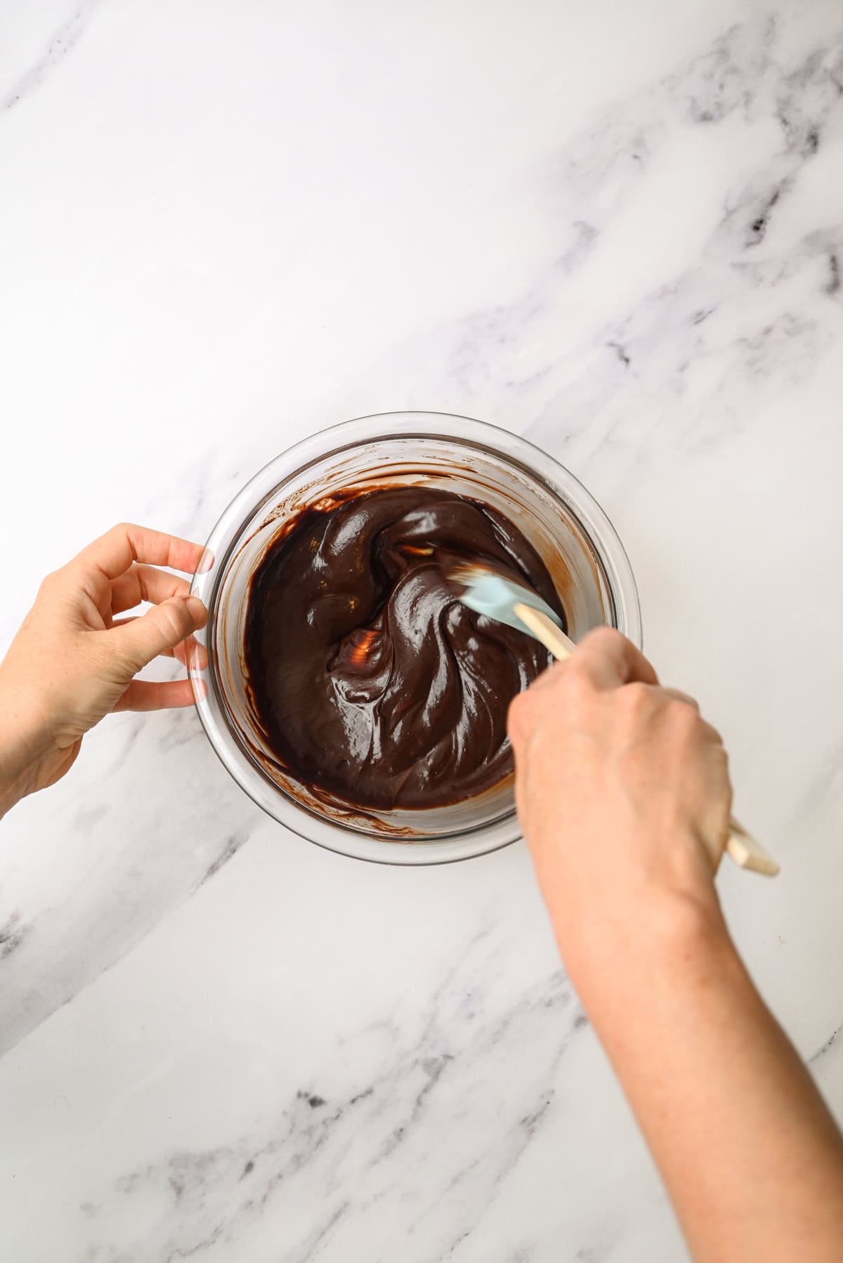 Two hands are mixing smooth, dark chocolate batter in a glass bowl with a spatula on a white marble surface.