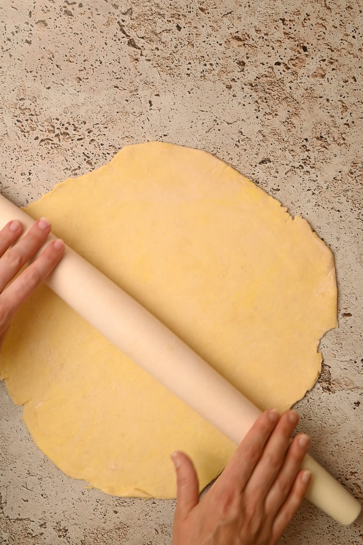 Two hands use a rolling pin to flatten a sheet of dough on a textured countertop.