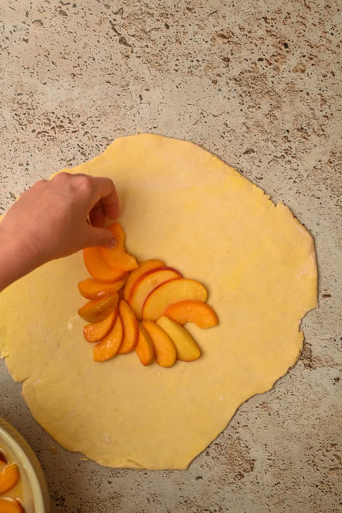 A hand arranges thin slices of peach on a sheet of rolled-out dough, preparing to make a fruit-filled pastry on a textured, light-colored surface.
