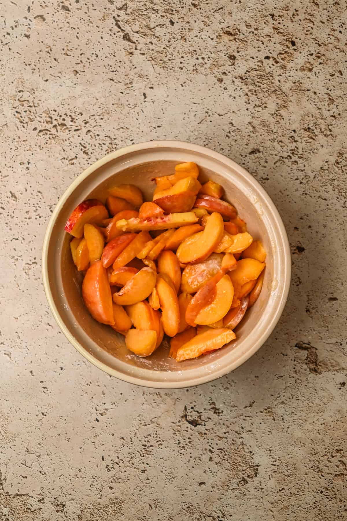 A beige bowl filled with sliced fresh peaches sits on a mottled, tan countertop. The peach slices are vibrant orange and appear to be lightly tossed, possibly with sugar or another coating.
