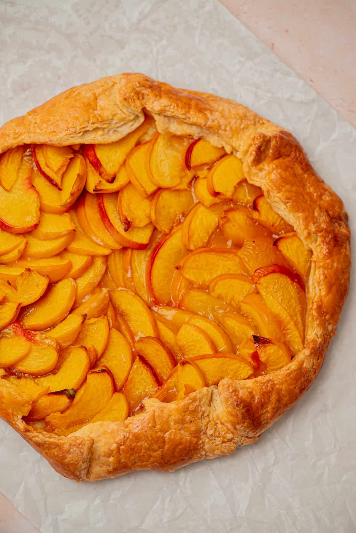 A rustic peach galette with golden, flaky crust and neatly arranged peach slices sits on crumpled parchment paper.