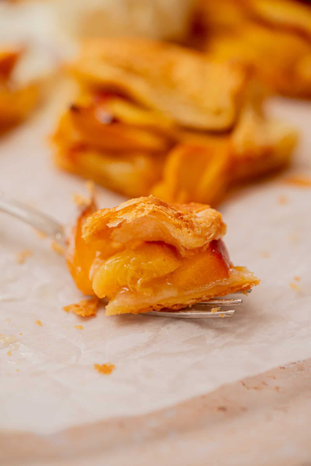 A close-up of a fork holding a bite of flaky peach pastry, with more pieces of the pastry blurred in the background on parchment paper.