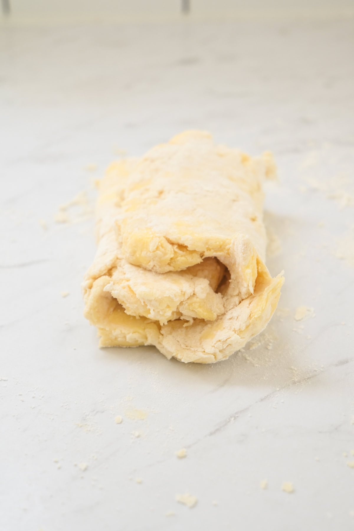 A piece of folded, flaky biscuit dough rests on a lightly floured, white marble surface, ready to be rolled out for baking.