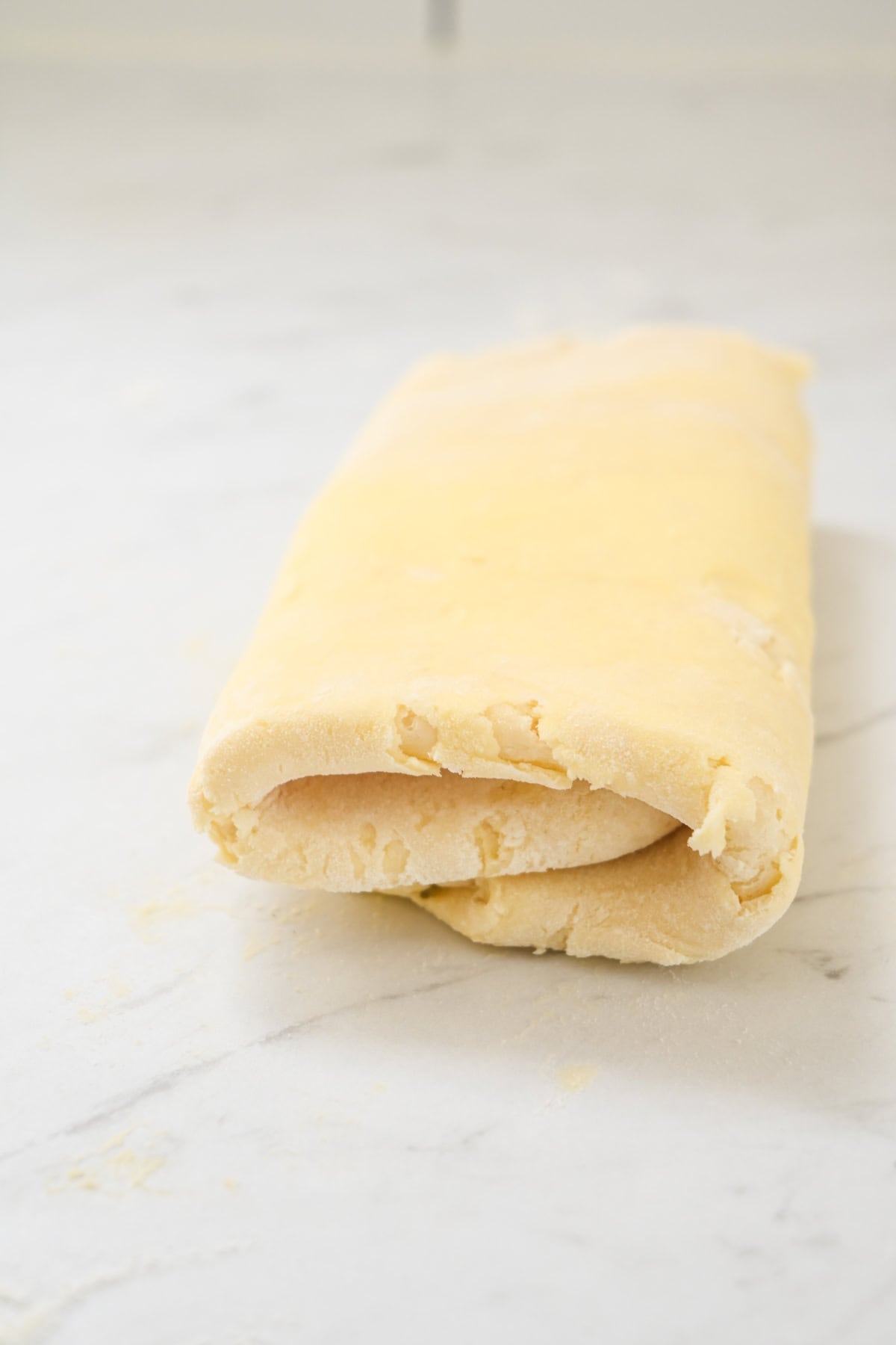 A folded piece of pale, uncooked pastry dough rests on a lightly floured marble surface.