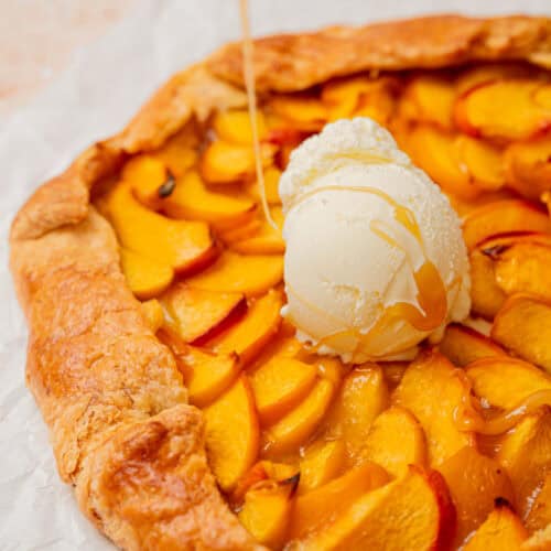 A rustic peach galette topped with a scoop of vanilla ice cream is being drizzled with honey. The galette’s golden, flaky crust frames vibrant peach slices on parchment paper.