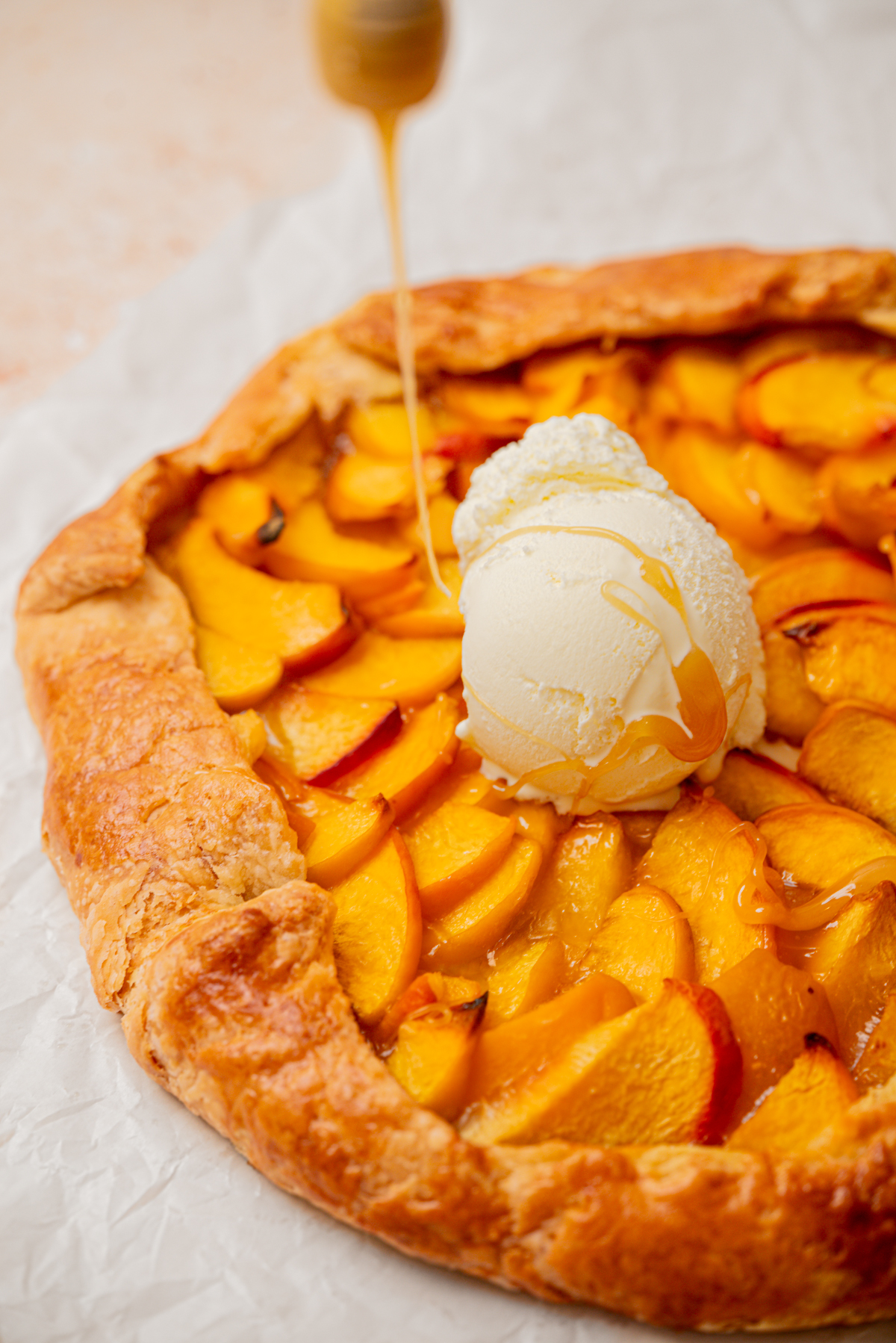 A rustic peach galette topped with a scoop of vanilla ice cream is being drizzled with honey. The galette’s golden, flaky crust frames vibrant peach slices on parchment paper.