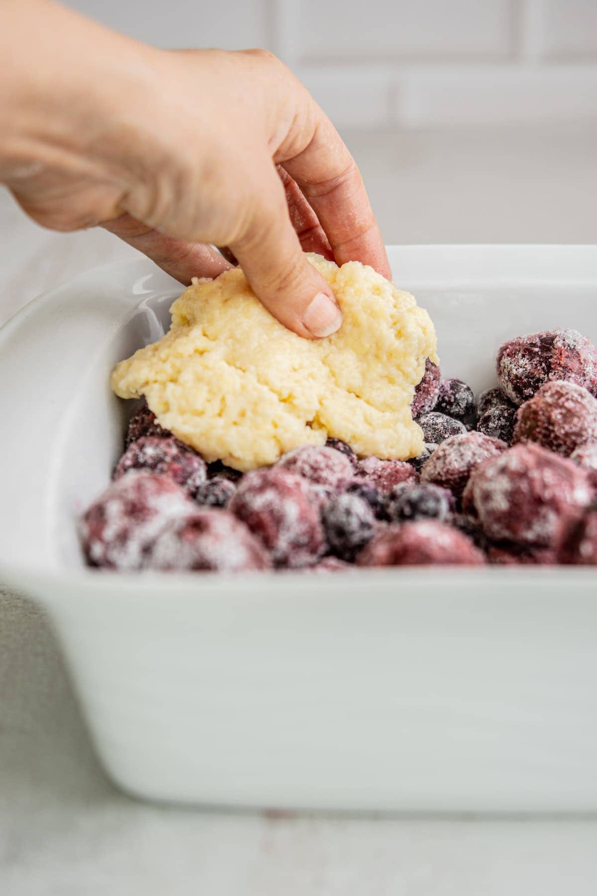 A hand places biscuit dough on top of sugared mixed berries in a white baking dish, preparing a cobbler dessert.