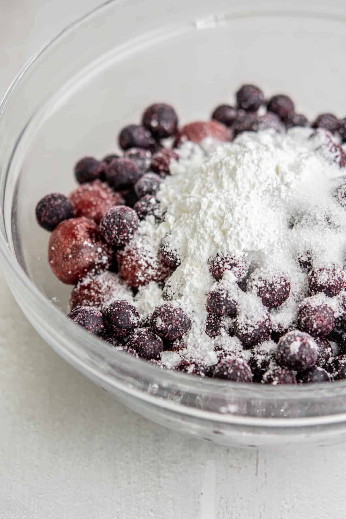 A glass bowl filled with frozen blueberries coated in a generous amount of white cornstarch or powdered sugar, sitting on a white surface.
