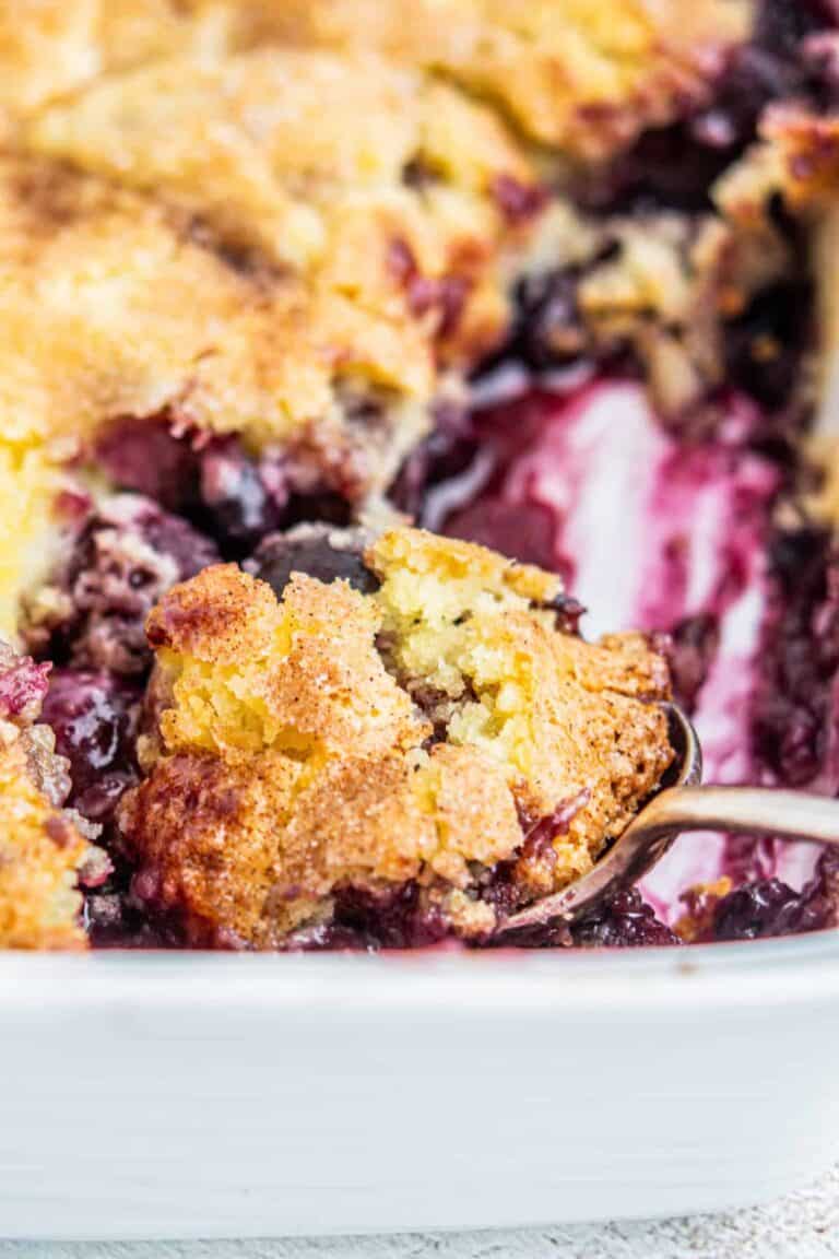 A close-up of a golden-brown blueberry cherry cobbler in a white baking dish, with a spoon lifting out a portion to reveal the juicy, purple berry filling underneath the crispy topping.