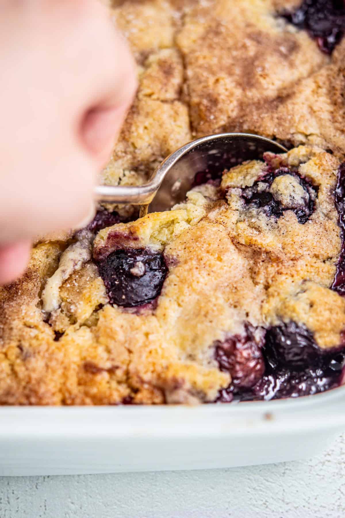 A close-up of a hand scooping out a portion of blueberry cobbler from a baking dish, showing golden, crumbly topping and bubbling blueberries.